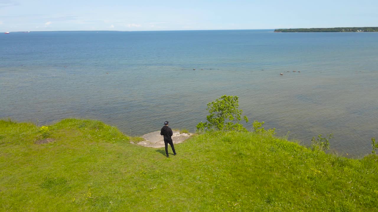 Aerial drone orbiting around a man with black clothing standing on a steep cliff bank at Baltic sea limestone shoreline during a summer sunny day with horizon visible in the distance. Vibrant colors.