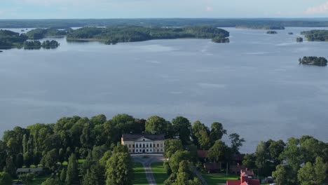 Backward-aerial-view-of-scenic-Taxinge-castle-by-trees-and-lake-in-Sweden