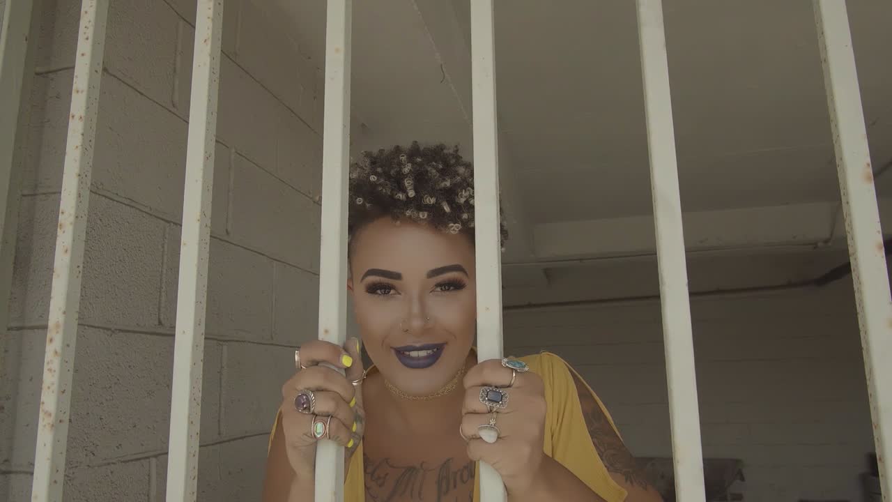 Woman with short hair in a yellow dress smiling and dancing behind some bars in a parking lot