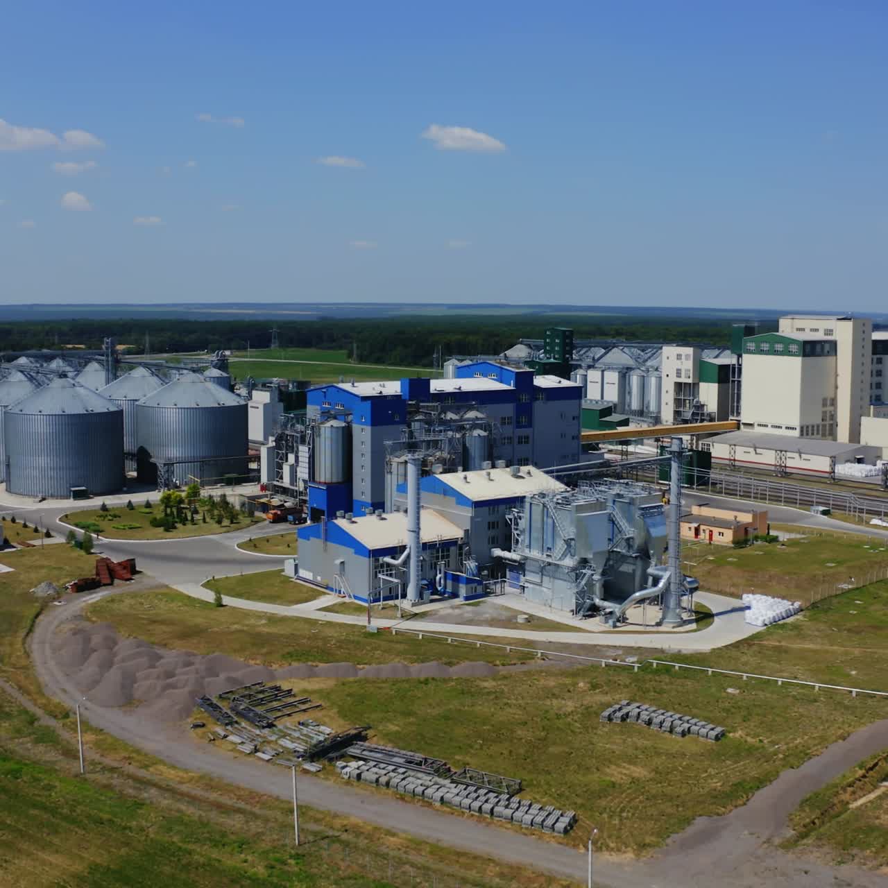 Agricultural industry. Modern factory on green field with steel elevators. Industrial buildings and granary under blue sky. Aerial view.