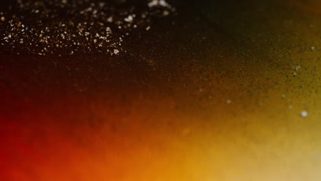 Macro view of shimmering gold particles swirling in brown, orange, and yellow ink forming abstract moving patterns