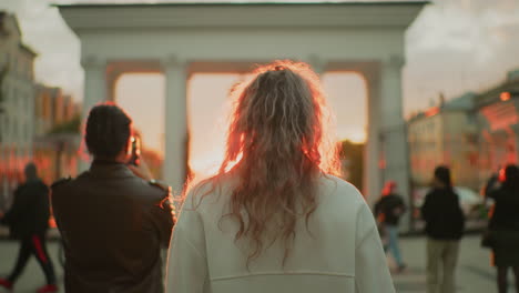 back view of woman with curly hair walking toward archway during sunset with strong red sunlight shining creating dramatic silhouette effect in busy urban environment