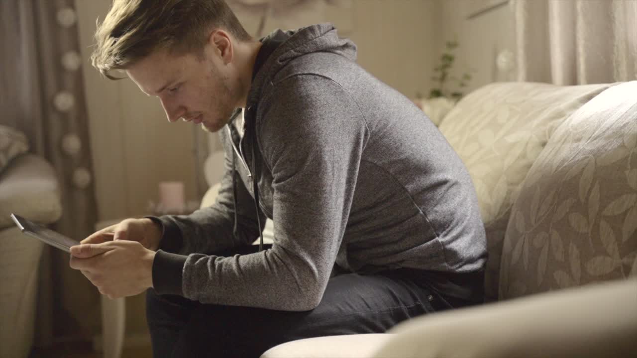 A male student sits on the sofa as he uses a portable tablet device to work