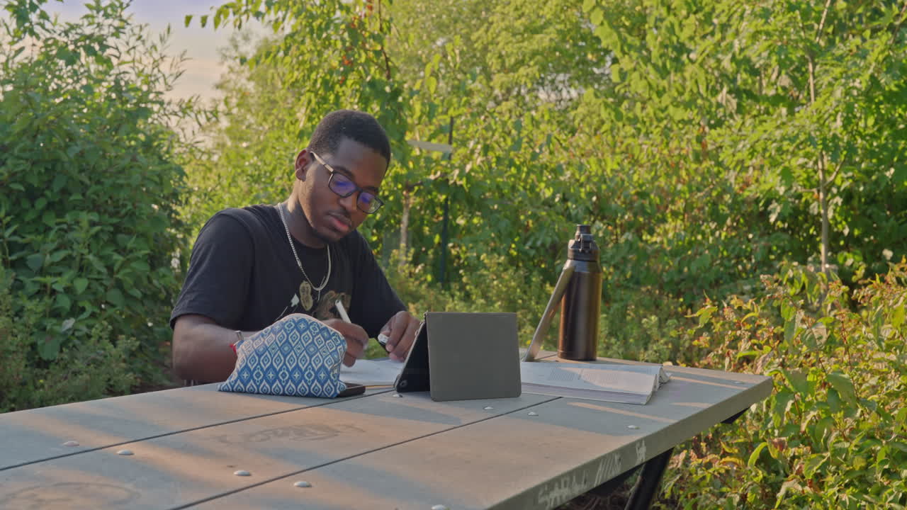 Young Black student sitting at a park table, using tablet, notebook and water bottle while studying outdoors. Dolly shot moving left, focus on education, technology, diversity and sustainability