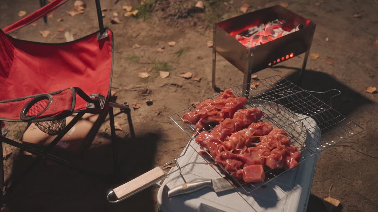 Panggangan berkemah di samping kursi merah di bawah cahaya senja yang redup, panggangan arang portabel dan tusuk sate daging marinasi di atas parutan kawat, bara api yang menyala dan aroma berasap, seorang pendaki menyiapkan barbekyu di meja lipat kecil.