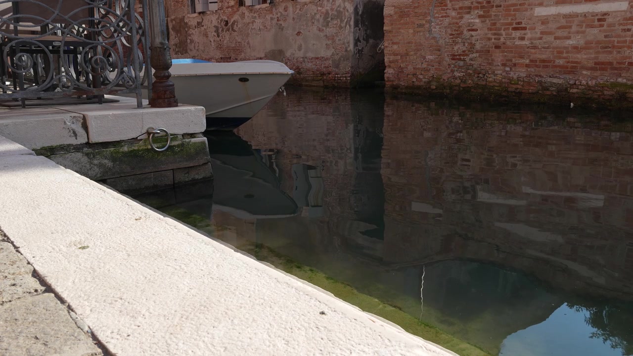 Vista statica di una barca ormeggiata su un tranquillo canale di Venezia con acqua calma che riflette storici edifici in mattoni in un'autentica ambientazione veneziana