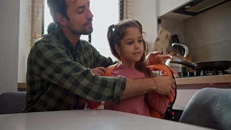 A-happy-brunette-man-in-a-green-checkered-shirt-helps-his-little-daughter,-a-brunette-girl-in-a-pink-dress,-put-an-orange-backpack-on-her-back-and-in-the-kitchen-in-a-modern-apartment