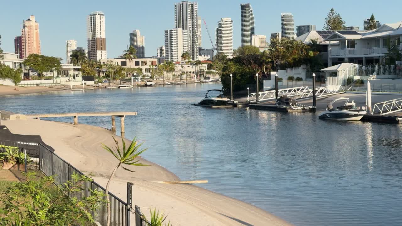 Silver gull flies above calm canal with luxury homes, boats, and city skyline in daylight