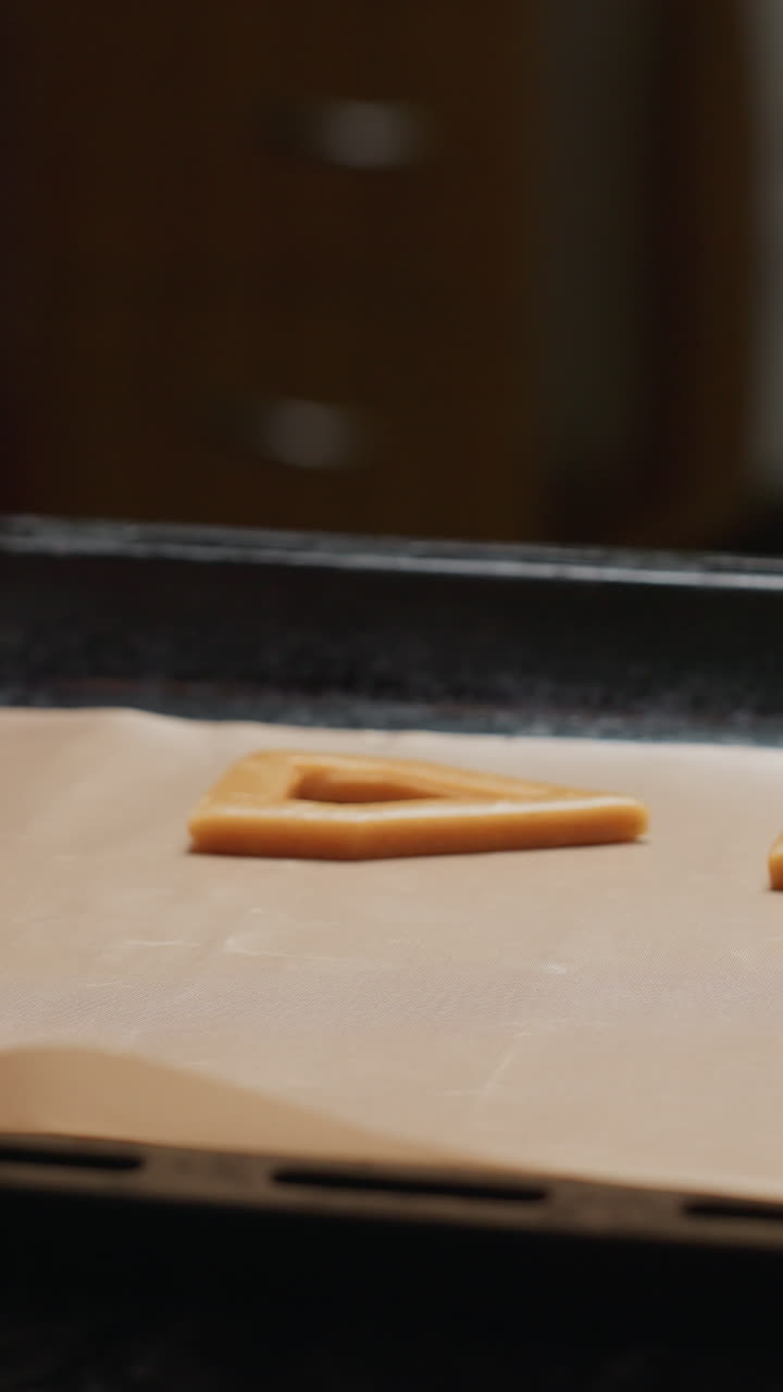 primo piano di un fornaio che mette l'impasto a forma di biscotto su carta pergamena in vassoio, che si prepara per la cottura, con rotoli, tagliatori di biscotti e altri strumenti che circondano l'impaeso in cucina a casa