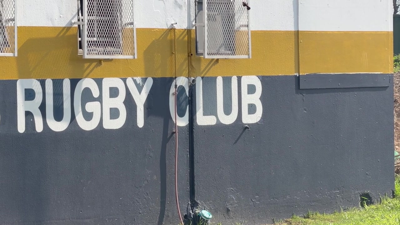 Close-up do exterior de um edifício de clube de rugby, apresentando letras brancas e ousadas “RUGBY CLUB” pintadas numa parede azul-marinho, com uma faixa amarela mostarda e uma seção superior branca