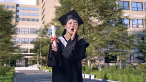 asian woman student graduates in cap and gown holding diploma and yelling positively with hand over mouth in front of a magnificent university building