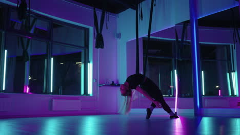 a woman does sports yoga in the air on a hammock in neon light performing graceful exercises and movements from stretching and dancing in slow motion.