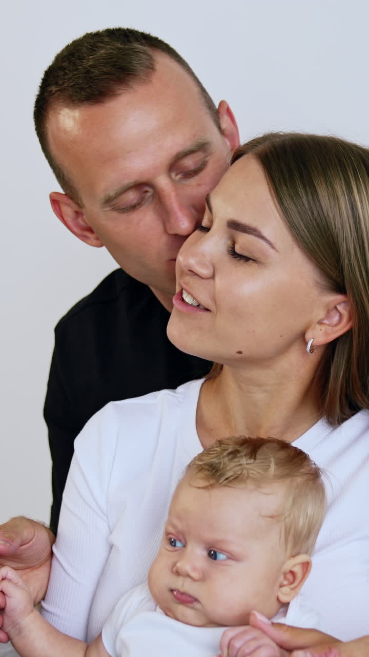 Marito bacia la moglie che tiene in braccio un bambino biondo. Famiglia felice di tre persone su sfondo bianco. Video verticale.