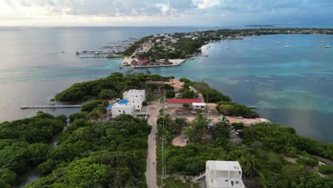 Drone-view-in-Belize-flying-over-caribbean-dark-and-light-blue-sea,-a-white-sand-caye-covered-with-palm-trees-and-restaurants-on-a-cloudy-day