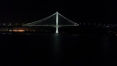 Toma-Aérea-De-Vehículos-Que-Se-Desplazan-Por-El-Puente-De-La-Bahía-De-San-Francisco–oakland-Con-La-Ciudad-De-Fondo-Por-La-Noche