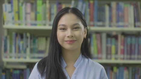 Joven-Estudiante-Universitaria-Sonriendo-A-La-Cámara-Con-Libros-De-La-Biblioteca-En-El-Fondo