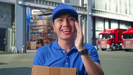 Close Up Of Asian male Courier In Blue Uniform Smiling And Talking To Camera While Delivering A Carton, Outside of Logistics Distributions Warehouse