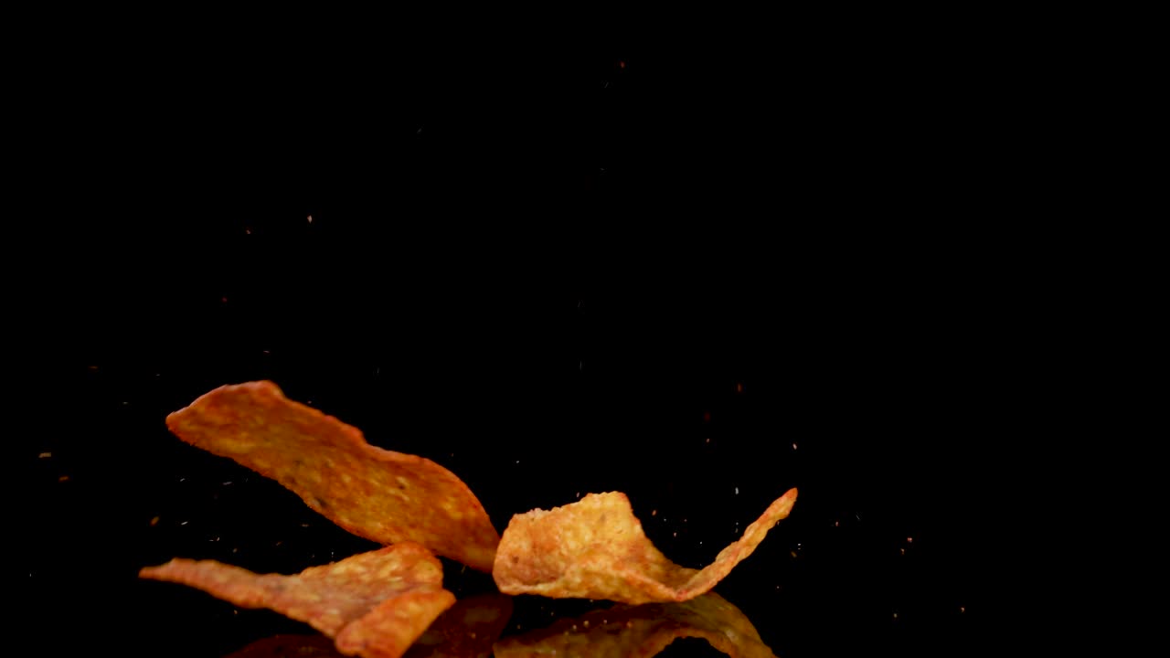 Crispy tortilla chips cascade and land on a dark surface, illuminated by dramatic studio lighting