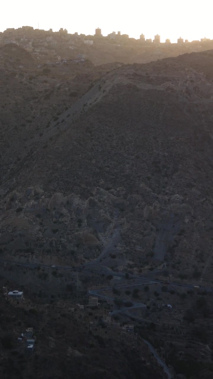Aerial view slowly revealing villages nestled in the rugged, mountainous landscape of Yafa'a, yemen, with the warm light of sunrise illuminating a distant city skyline on the horizon. In Yafa'a, Yemen