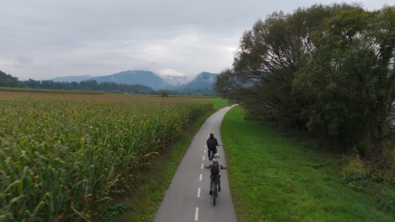 Aerial: couple riding e-bikes in the countryside along Drava River with moody sky during the day, tracking drone shot