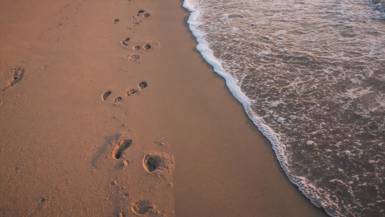 Onde che si infrangono sulla sabbia bagnata vicino a impronte umane sulla spiaggia, con la marea che cancella delicatamente le impronte