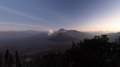 Puffing-smoking-Mount-Bromo-Volcano-at-sunset-twilight-with-motion-control-moving-thistle-flower-illuminated-foreground