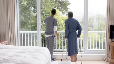 diverse male doctor helping senior male patient using walking frame at home, slow motion