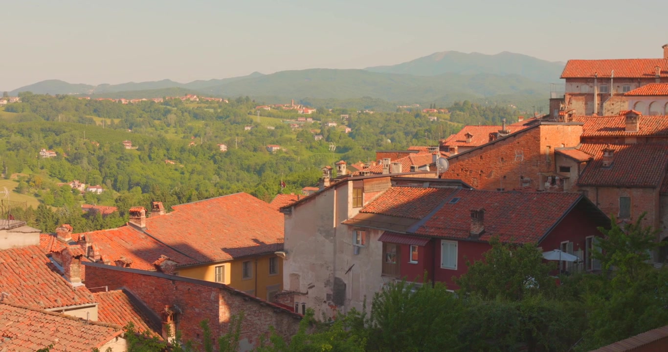 Vista dall'alto sui tetti tradizionali in tegole di terracotta di case storiche, verso dolci colline verdi e montagne distanti, sotto un cielo caldo e soleggiato in Piemonte, Italia.