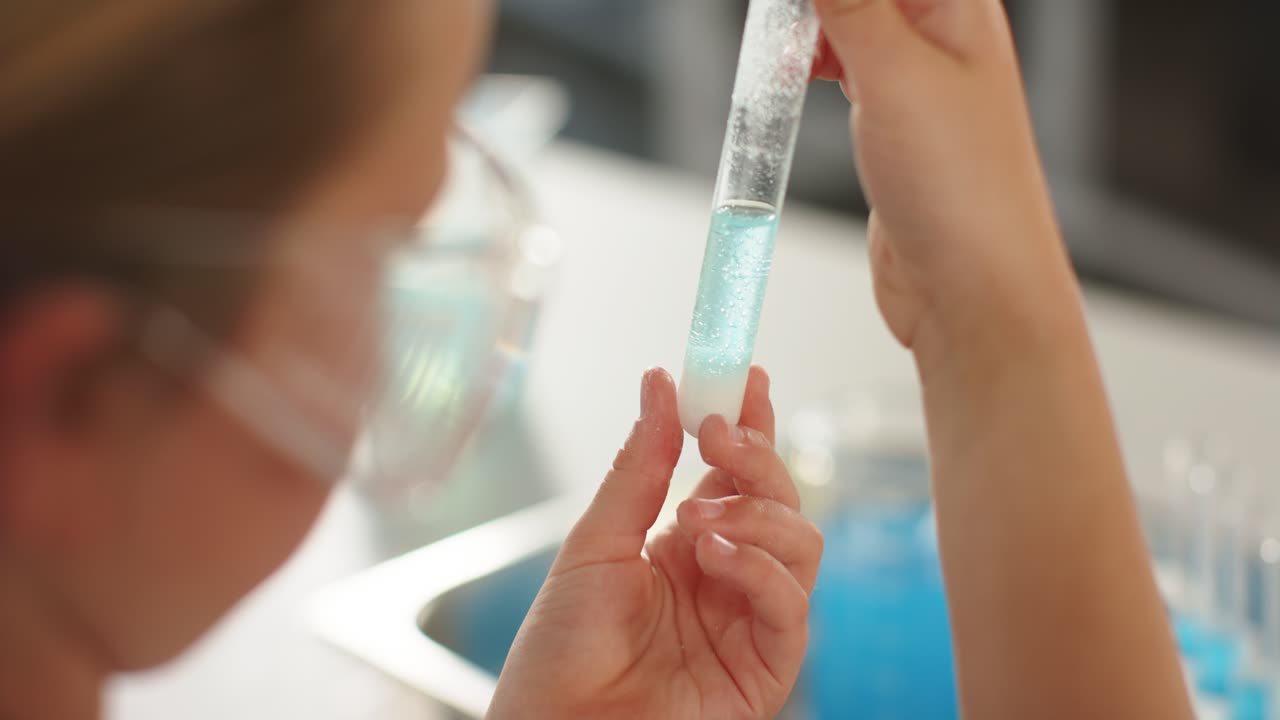 Youth female in goggles seeing sediment raising test tube at bench inspecting turquoise liquid