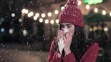 vista de cerca de una mujer caucásica enferma con abrigo rojo sonándose la nariz en la calle mientras nieva en navidad