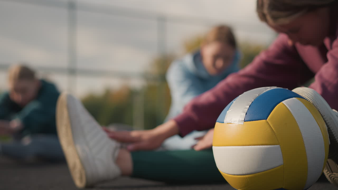 근처에 배구 공을 들고 스트레칭하는 여자의 클로즈업, 다른 여자들이 야외 스포츠 경기장에서 배경에서 운동하면서 활발한 피트니스 훈련 분위기를 만니다.