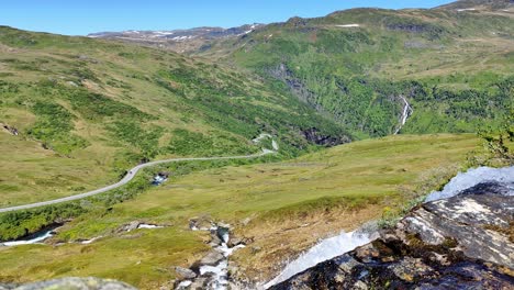 Wild-nature-with-road-rv13-crossing-mountain-Vikafjell-in-Norway-seen-from-idyllic-mountainside-with-a-fresh-crisp-river-stream-passing-in-foreground-close-to-camera---Static