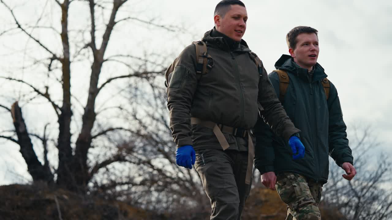 Side view of two confident young men in dark green army uniforms and blue medical gloves walking along the military training ground and combat practice and communicating with backpacks on their backs