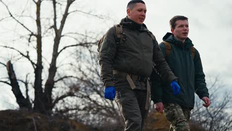 Vista-Lateral-De-Dos-Jóvenes-Confiados-Con-Uniformes-Militares-Verde-Oscuro-Y-Guantes-Médicos-Azules-Caminando-Por-El-Campo-De-Entrenamiento-Militar-Y-Prácticas-De-Combate-Y-Comunicándose-Con-Mochilas-En-Sus-Espaldas.