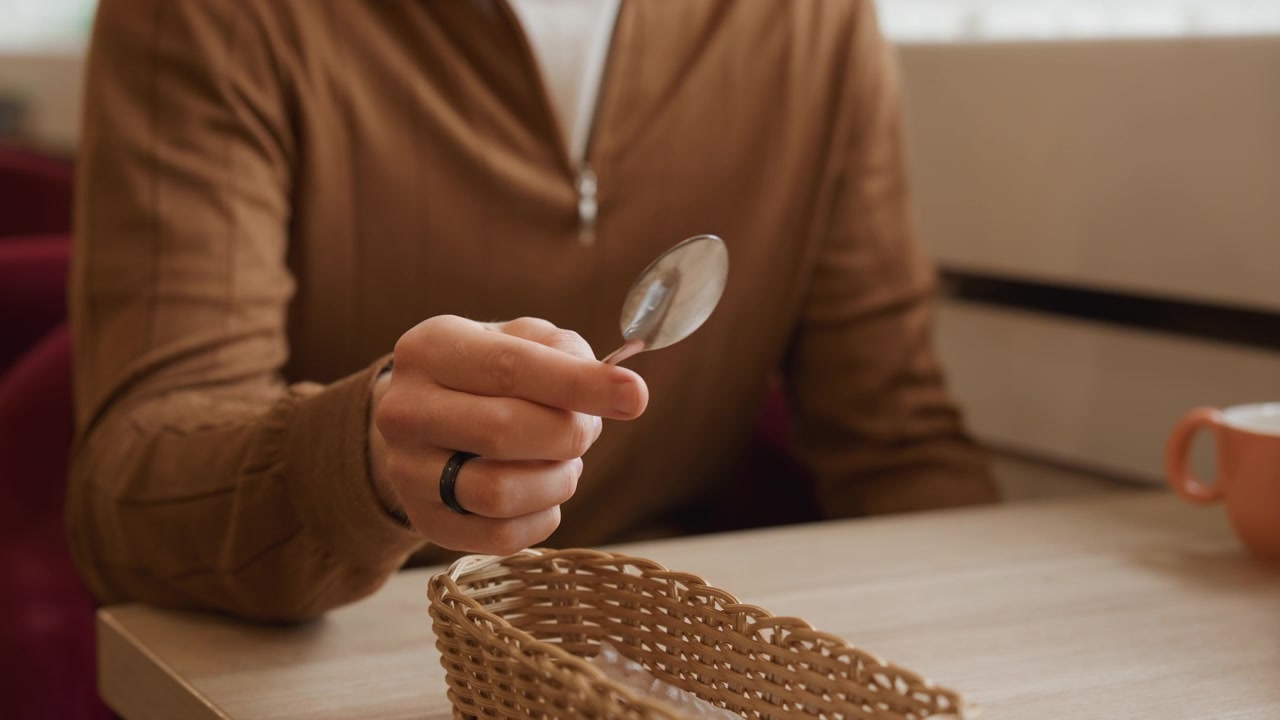Primo piano di un uomo che sistema le posate in un cesto intrecciato su un tavolo di legno con luce soffusa. L'uomo indossa una giacca marrone e ha le mani visibili con anelli, mentre una tazza arancione è vicina. Ambiente interno
