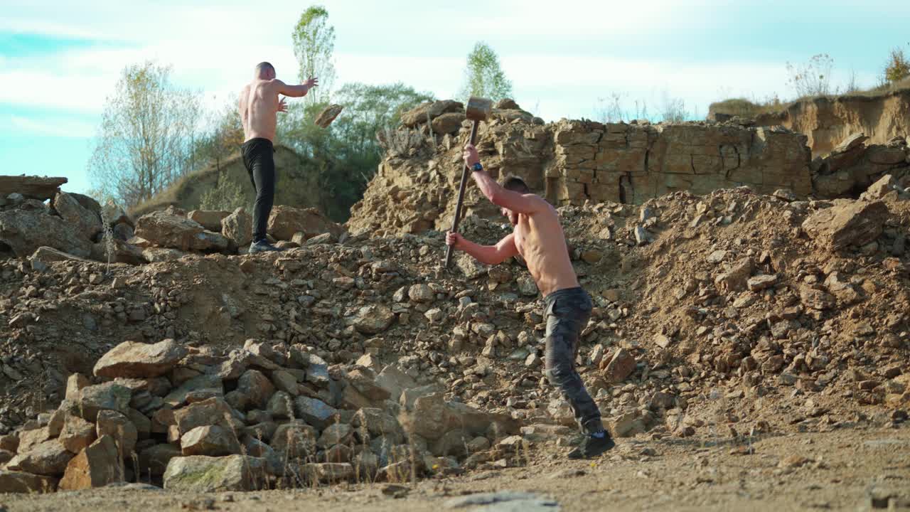 Two sportsmen breaking stones outdoors. Shirtless guy with a hammer on the rocky background. Athlete men training their body with hard exercises.