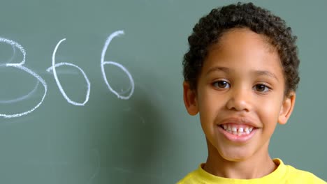 front view of african american schoolboy standing against green chalkboard in classroom at school 4k