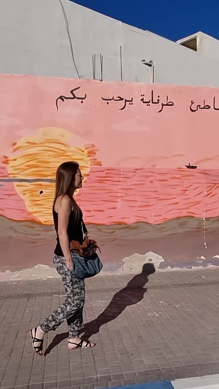 Vista laterale di una turista che cammina su un marciapiede a Tarfaya, Marocco. Un murale colorato che raffigura il deserto e il mare adorna la parete.
