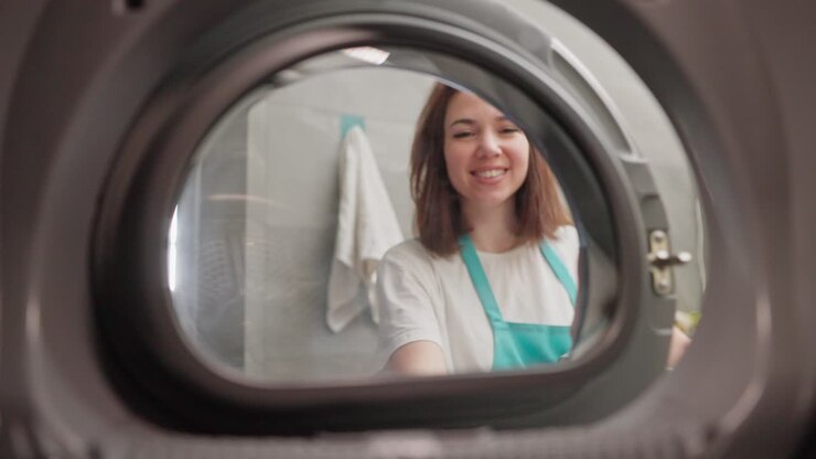 A happy brunette girl in a white T-shirt and a blue apron took out clean things from the washing machine and closes its door while cleaning the apartment on call