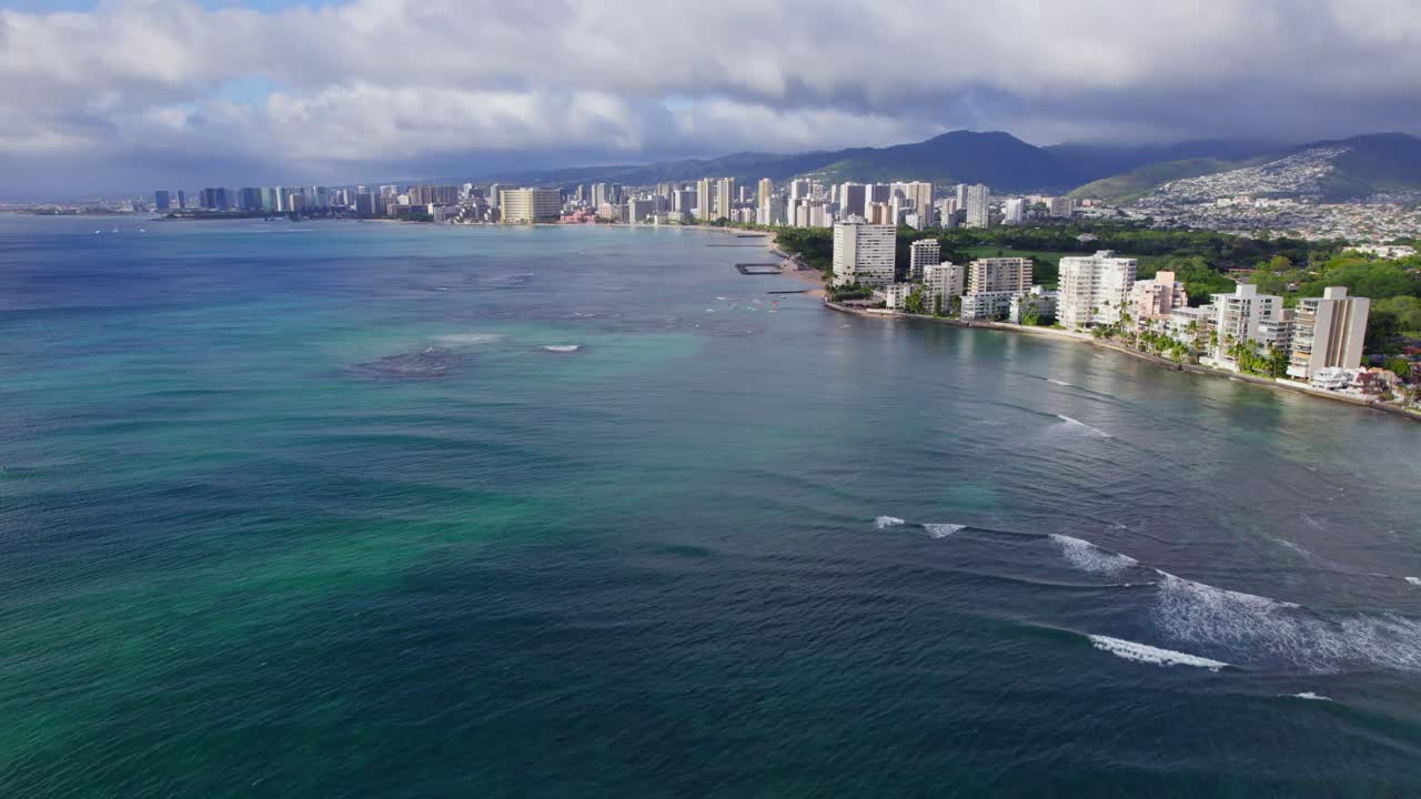 Riprese drone sopra l'acqua blu acquamarina dell'Oceano Pacifico lungo la costa dell'isola di Oahu, Hawaii. Lo skyline di Honolulu si estende lungo la costa con formazioni vulcaniche all'orizzonte
