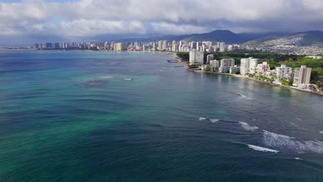 Imágenes-De-Drones-Sobre-El-Agua-Azul-Agua-Del-Océano-Pacífico-A-Lo-Largo-De-La-Costa-De-La-Isla-De-Oahu,-El-Paisaje-Urbano-De-Hawaii-Honolulu-Bordea-La-Costa-Con-Formaciones-Volcánicas-En-El-Horizonte