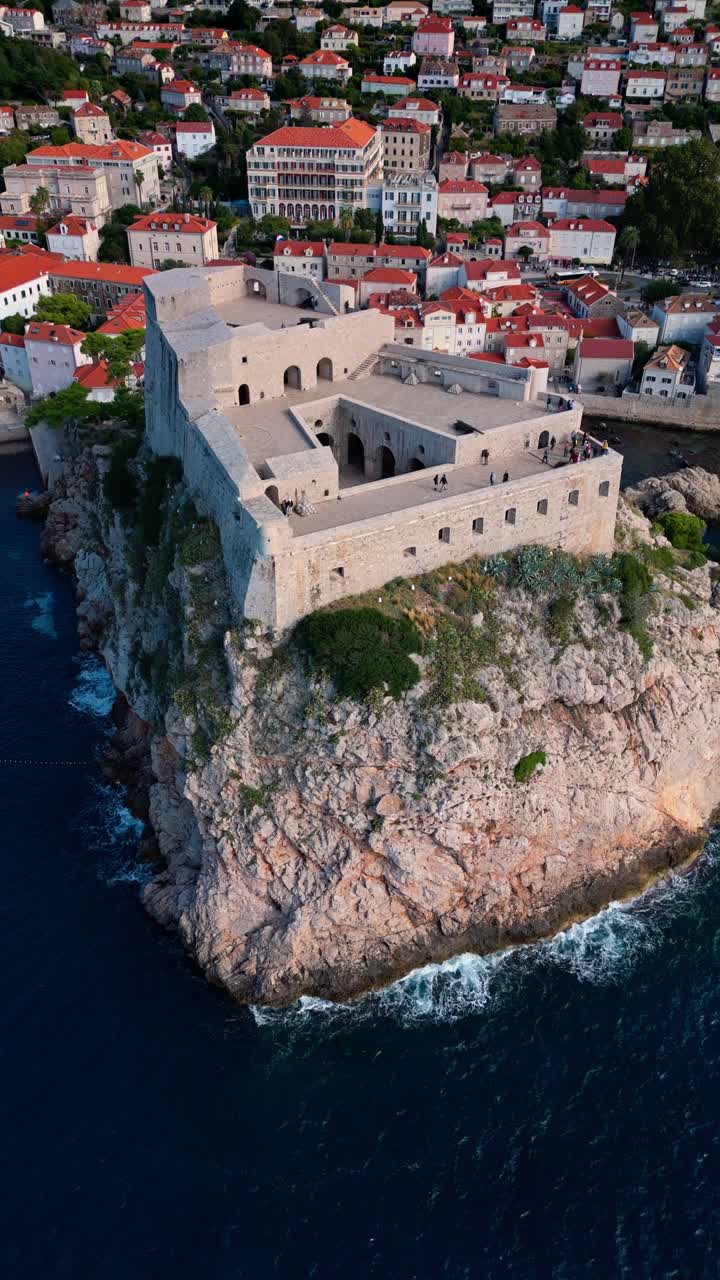 Aerial portrait drone shot flying down and tracking Fort Lovrjenac on the Adriatic Sea cliff coast, capturing the fortress perched atop rugged cliffs with deep blue waters and rocky shoreline below