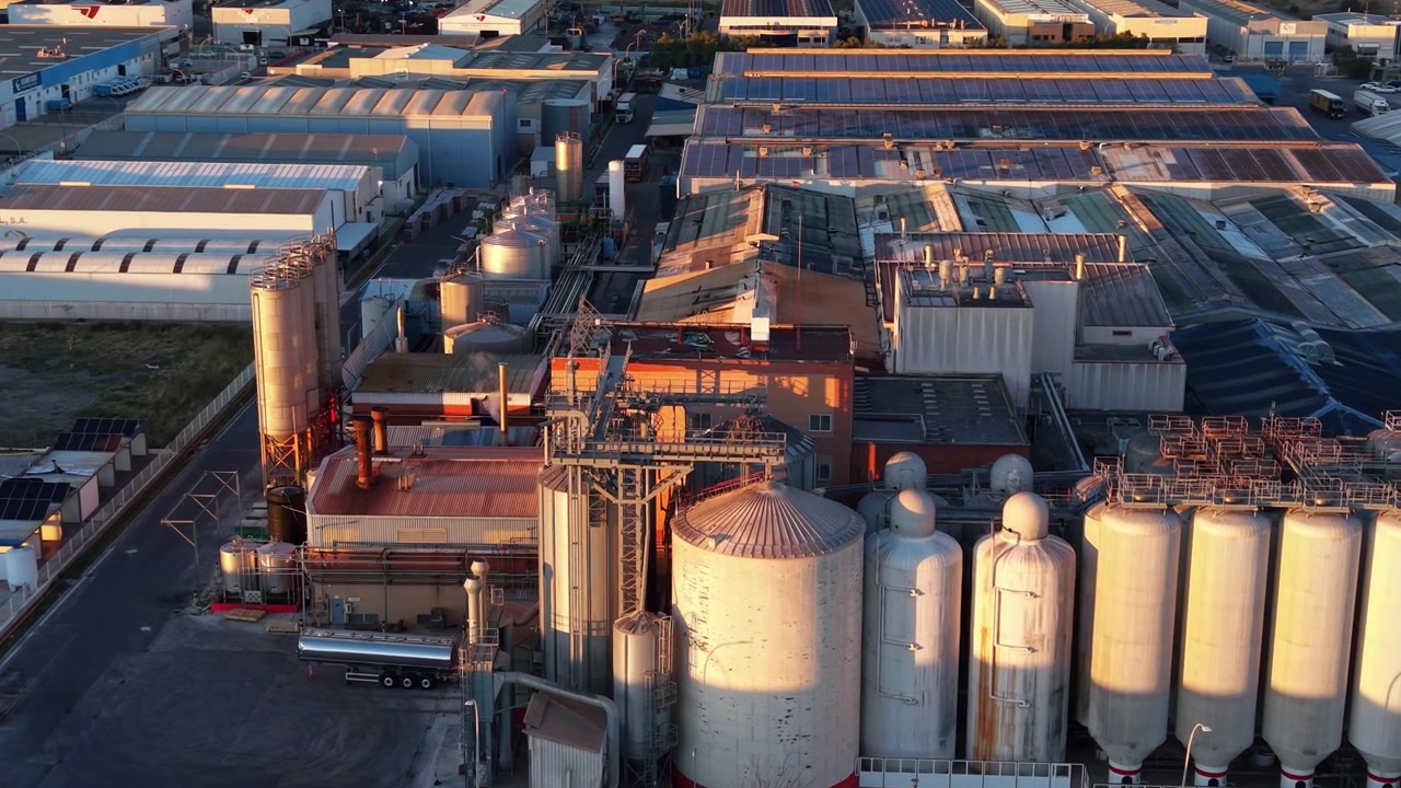 Overhead view of a large industrial complex at golden hour featuring grain silos, storage tanks, factory warehouses with solar panels and a tanker truck
