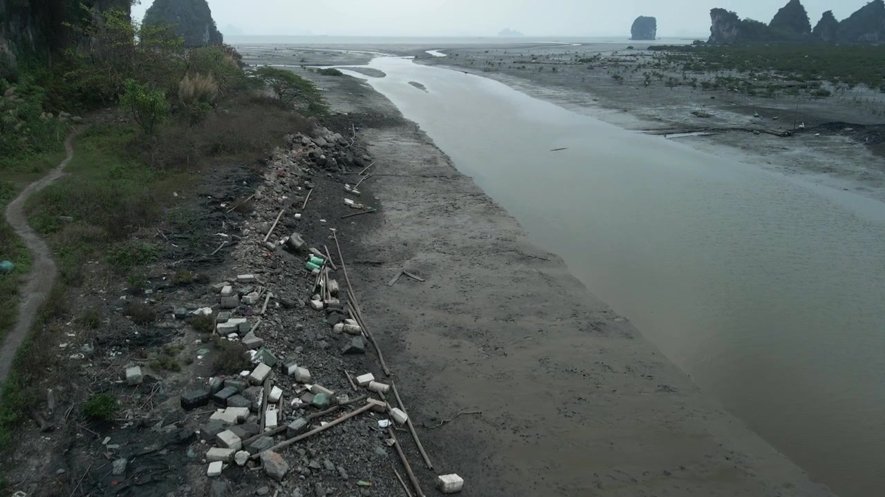 Fisherman's rubbish washed up along muddy banks of Halong Bay, Vietnam. Aerial drone shot along shoreline tilting up to reveal the waste disgarded and strewn along the waterline.