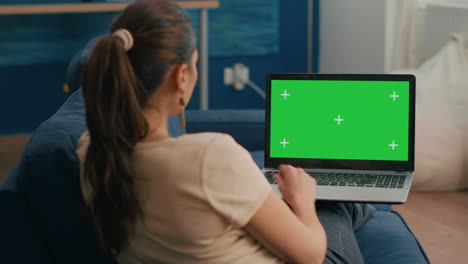 mujer freelancer sentada en el sofá en la oficina de casa escribiendo en una computadora portátil con una maqueta de pantalla verde clave de croma