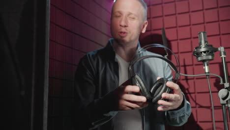 close-up of a man in black passing headphones to another man in a blue long-sleeve shirt against a vibrant red soundproof studio background, capturing a moment of preparation for a music session