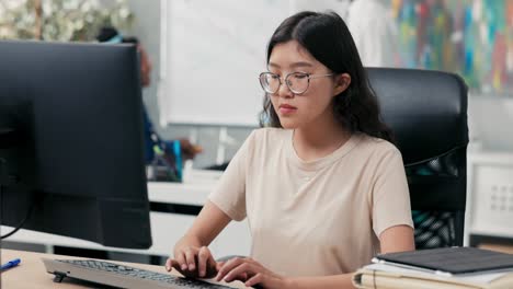 nerd girl in big glasses and tshirt sits in comfortable chair in front of desk, taps fingers on computer keyboard, sends messages, emails, writes back, runs company website, office life, corporate