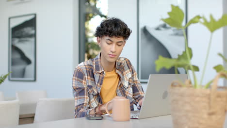 using laptop and smartphone, teenage boy working from home with coffee mug