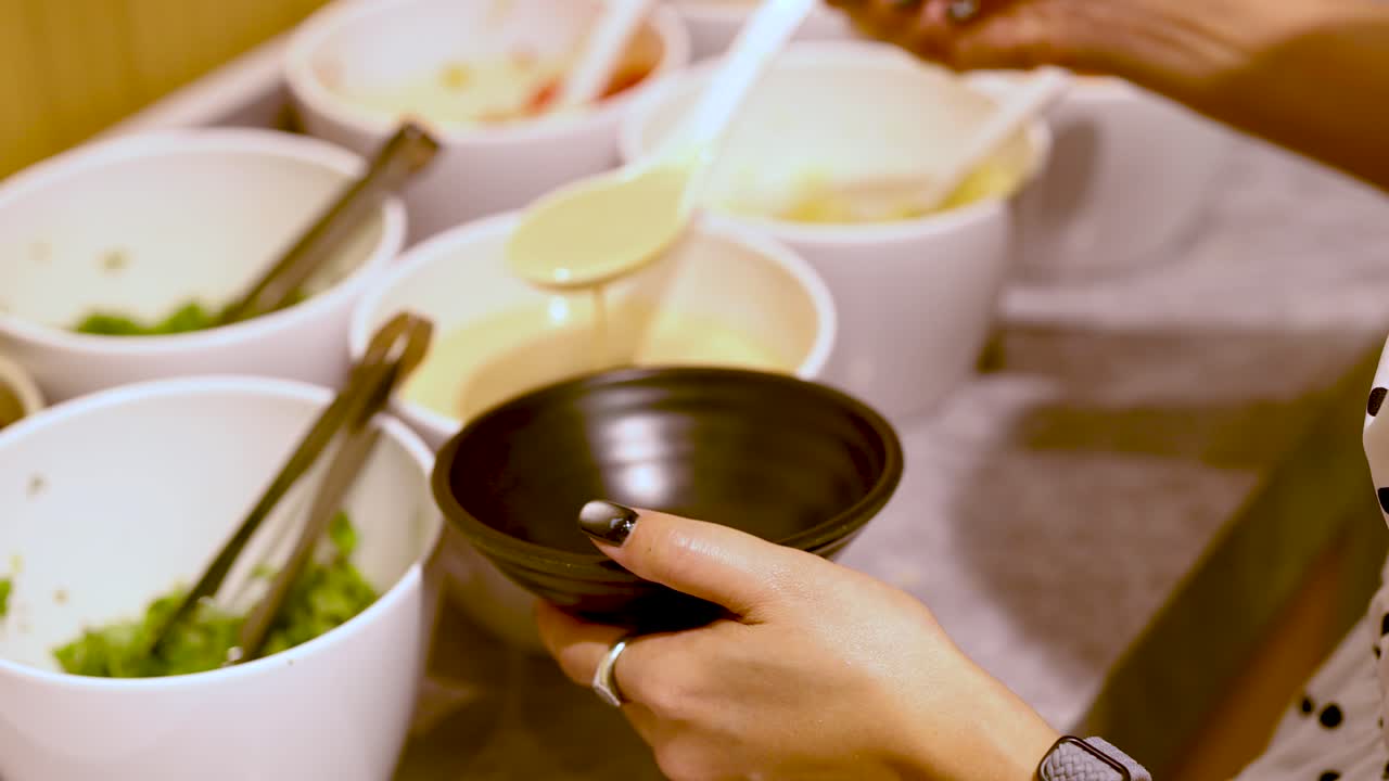 Person ladles creamy sesame sauce into bowl at well-lit hotpot condiment self-service station