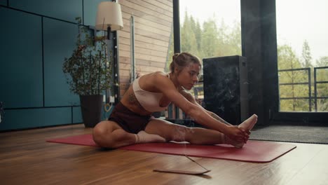 A-blonde-girl-in-a-white-top-stretches-on-a-special-rug-in-an-industrial-house-overlooking-a-coniferous-forest.-Near-her-smoking-incense